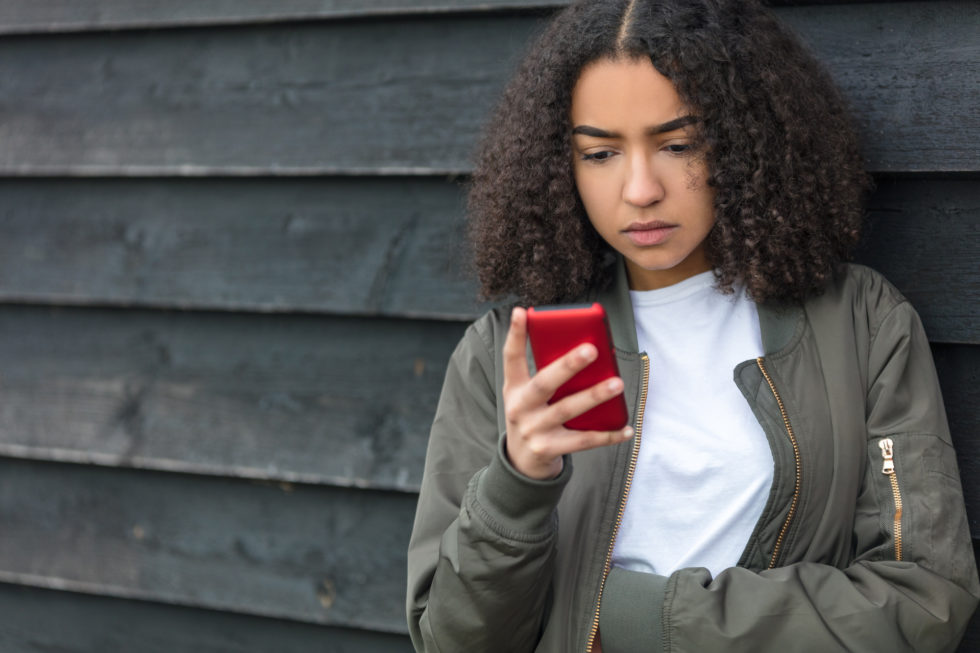 Outdoor portrait of beautiful sad depressed mixed race African American girl teenager female young woman texting or using social media on red cell phone wearing green bomber jacket